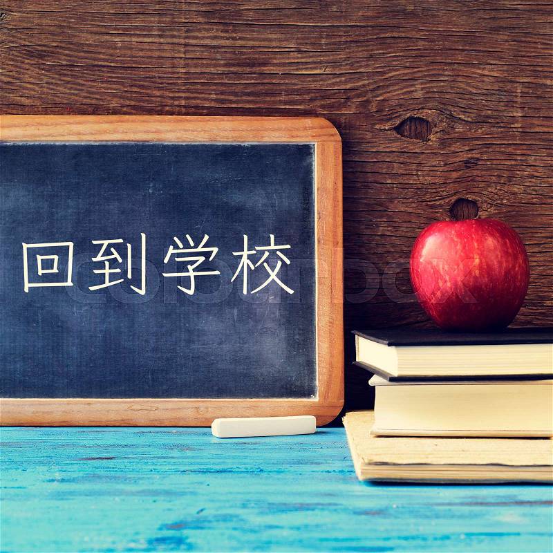 A chalkboard with the text back to school written in Chinese, a piece of chalk and an apple on a pile of books, placed on a blue wooden desk, against a rustic wooden background, stock photo