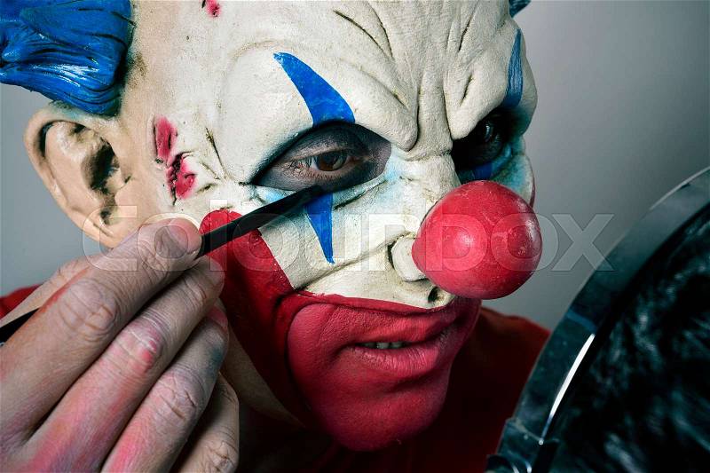 Closeup of a young man making up himself as an evil clown, using a mirror, stock photo