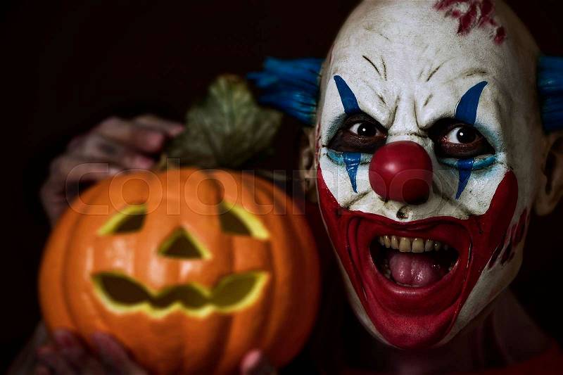 Closeup of a scary evil clown holding a carved pumpkin next to his head, stock photo