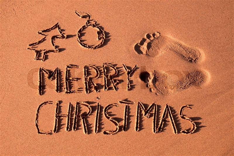 Some foot prints, the text merry christmas and the sketch of a christmas tree and a christmas ball carved in the sand of a beach, stock photo