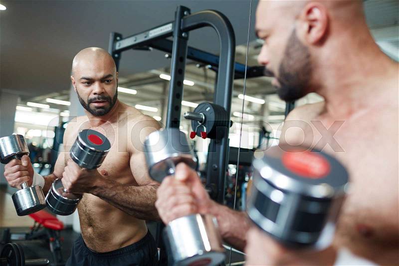 Portrait of shirtless muscular man looking in mirror, doing arm exercise working out with dumbbells during strength training in modern gym, stock photo