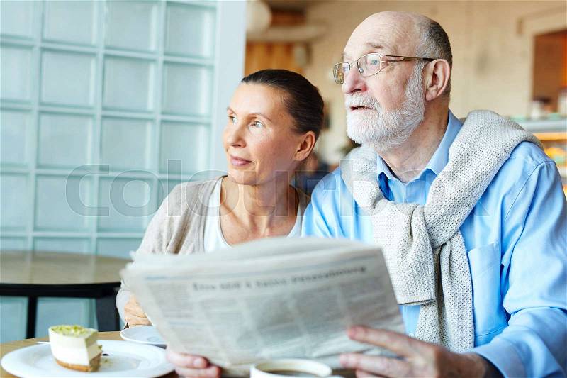 Modern couple of seniors with newspaper sitting in cafeteria, stock photo