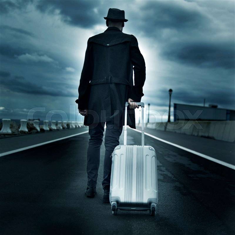 Closeup of a young man seen from behind carrying his rolling suitcase, with a dramatic effect, stock photo