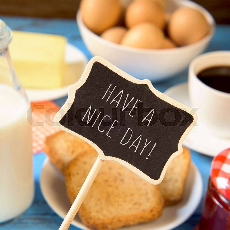 A chalkboard with the text have a nice day and a blue wooden table with a bottle of milk, a cup of coffee, some toasts in a plate, a jar of jam and a bar of butter, stock photo
