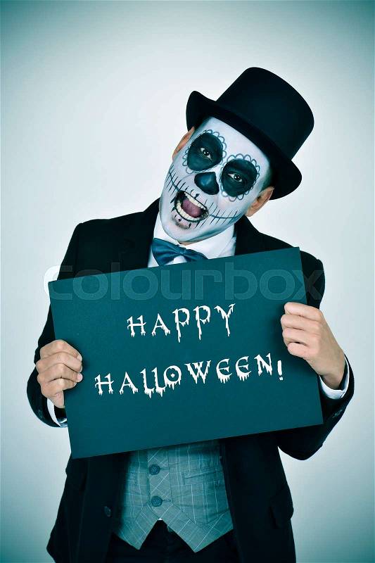 A man with mexican calaveras makeup, wearing jacket, vest, bow tie and top hat, shows a signboard with the text happy halloween, stock photo
