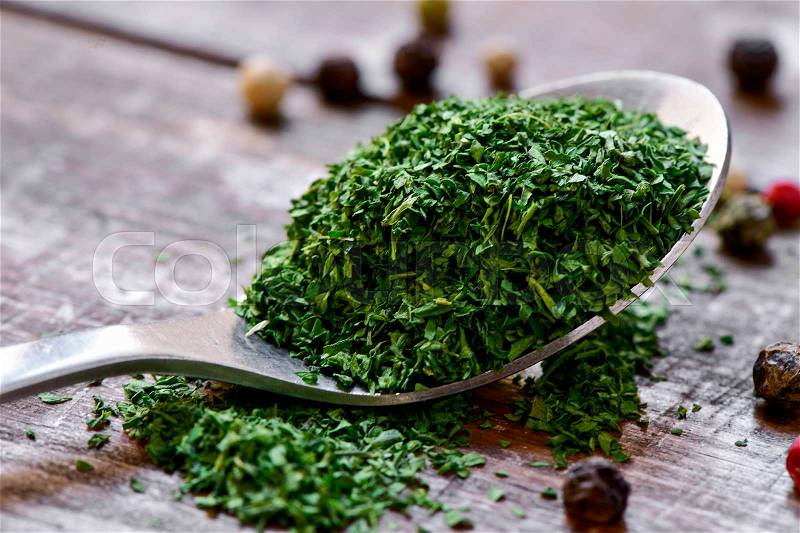 Closeup of a spoon with chopped dried parsley on a rustic wooden table with some peppercorns, stock photo