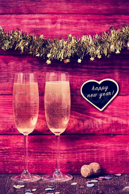 Confetti, a pair of glasses with champagne and a heart-shaped chalkboard with the text happy new year on a red rustic wooden background ornamented with tinsel, stock photo