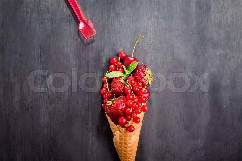 Fresh red summer berries - strawberries, red currants, cherries in the waffle cone in the form of ice cream and little spoon on the black wooden background. Healthy dessert. Top view. Space for text, stock photo