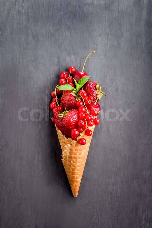 Fresh red summer berries - strawberries, red currants, cherries i with mint leaf in the waffle cone in the form of ice cream on the black wooden background. Healthy dessert. Top view. Space for text, stock photo