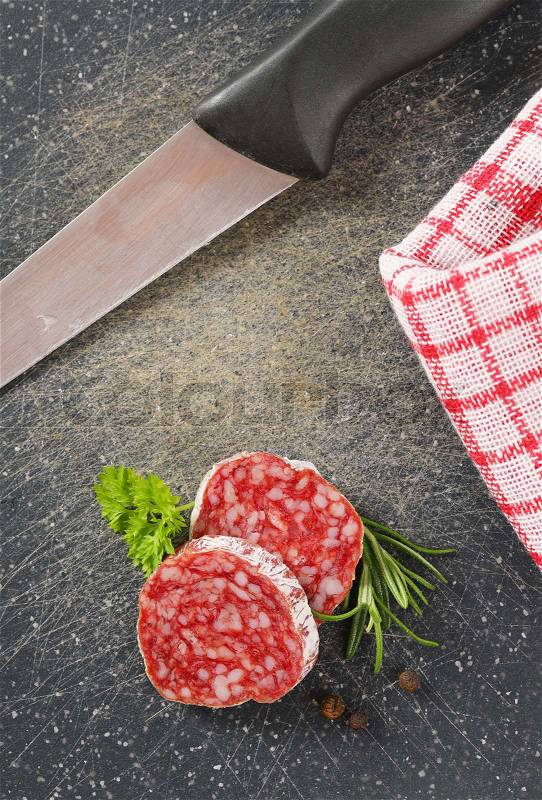 Slices of dry cured sausage with herbs and peppercorns on black cutting board, stock photo