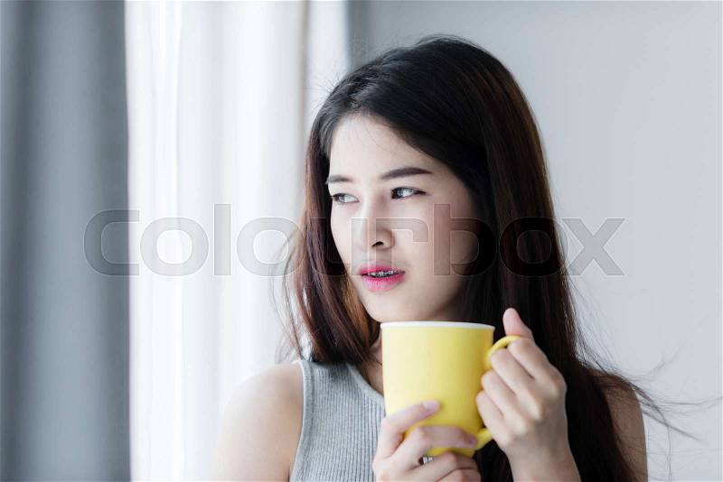 Asian woman holding yellow mug near window with natural light, stock photo