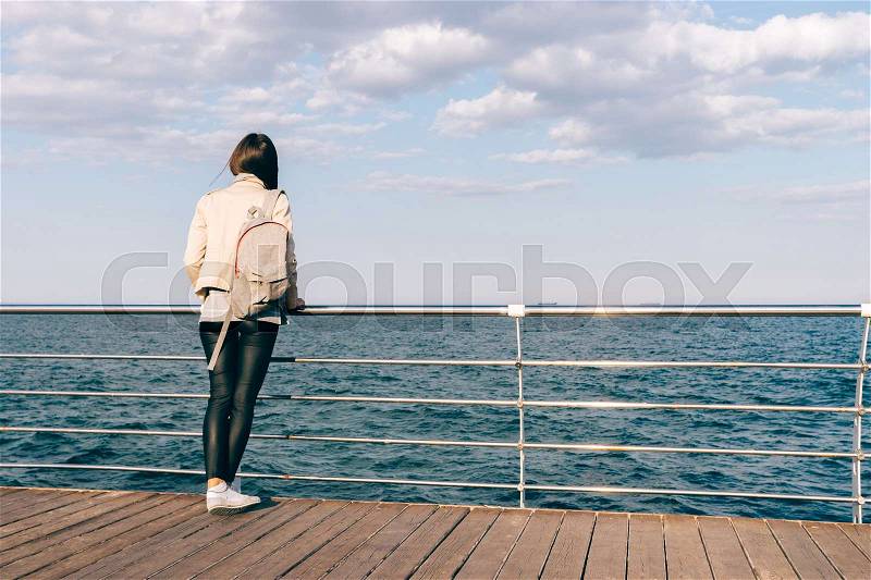 Woman in a beige jacket and black pants is standing on the waterfront and looking at the sea, stock photo