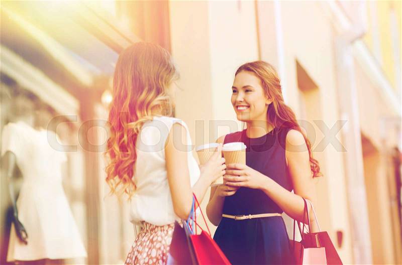 Sale, consumerism and people concept - happy young women with shopping bags and coffee paper cups talking at shop window in city, stock photo