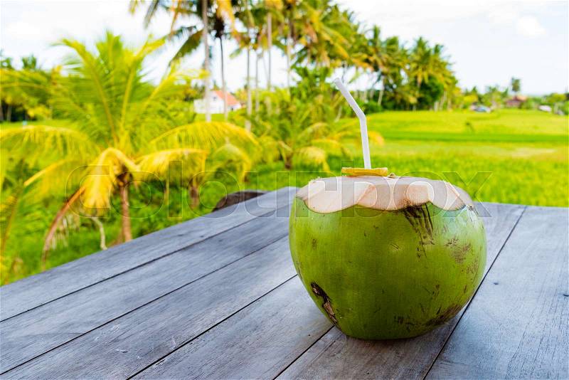 Green young coconut with straw on table, rice fields and coconut trees in backgroud. Copy space available, stock photo