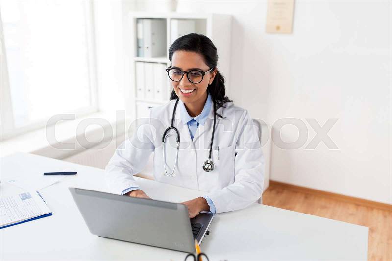 Healthcare, technology people and medicine concept - happy smiling female doctor in white coat and glasses typing on laptop computer at hospital, stock photo