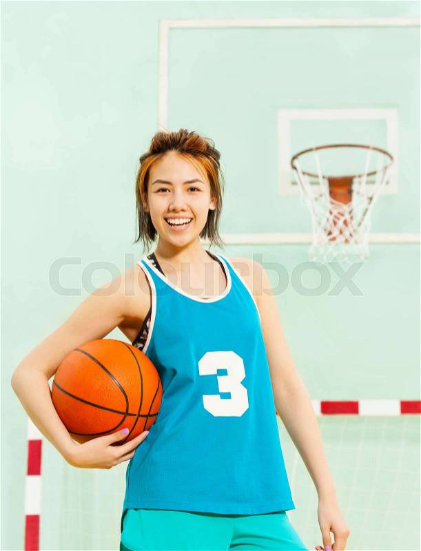 Portrait of happy teenage girl in uniform, basketball player, standing in gymnasium and holding ball, stock photo