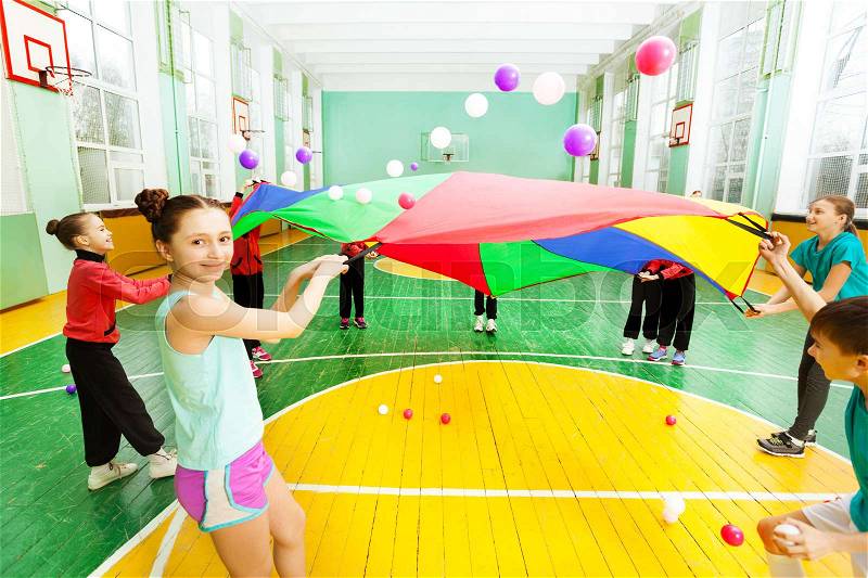 Portrait of preteen girl, waving rainbow parachute full of colorful balls, with her friends in sports hall, stock photo
