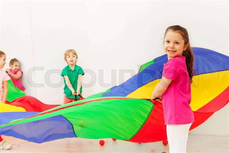 Portrait of happy five years old girl playing colorful parachute with her friends in gym, stock photo