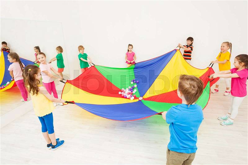 Happy children gathering around the party-coloured parachute and playing games in gym, stock photo