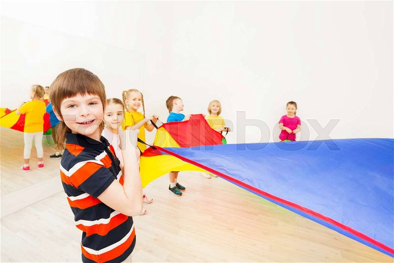 Portrait of smiling kid boy playing colorful parachute with friends in gym, stock photo