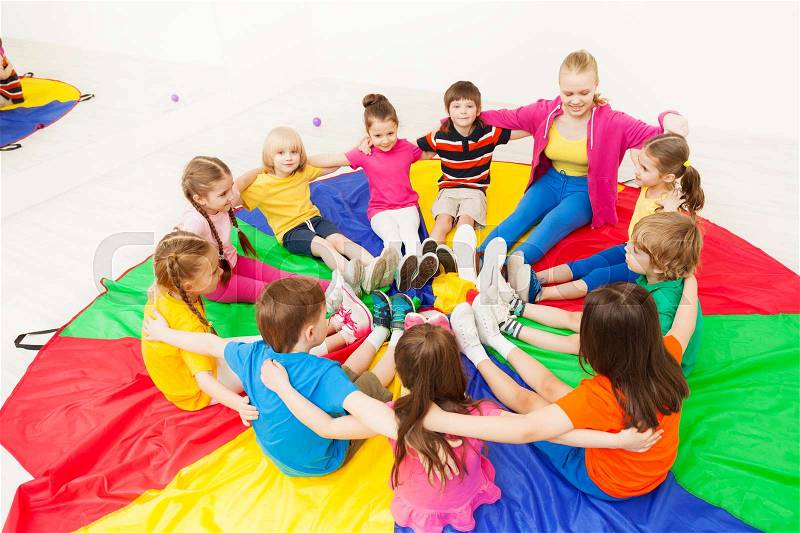 Big group of happy children sitting in a circle with nursery teacher, hugging and playing games, stock photo