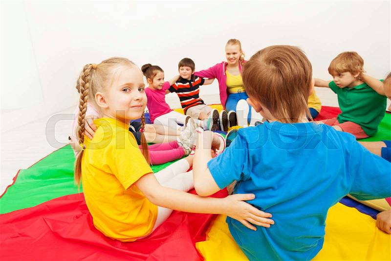Portrait of blond preschool girl playing games with friends, hugging and sitting in circle on colorful parachute, stock photo