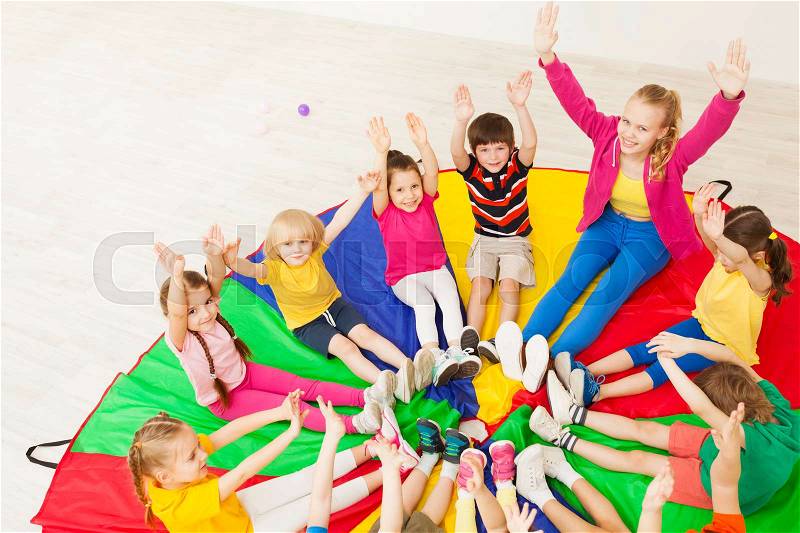 Portrait of young nursery teacher playing circle games with preschool children, sitting on rainbow parachute in gym, stock photo