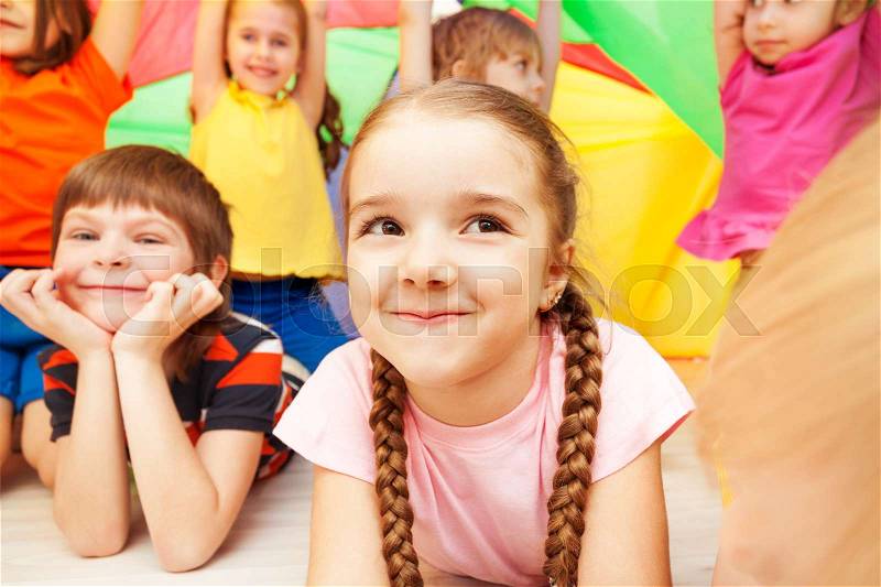 Close-up portrait of beautiful preschool girl playing with friends and hiding under rainbow parachute, stock photo
