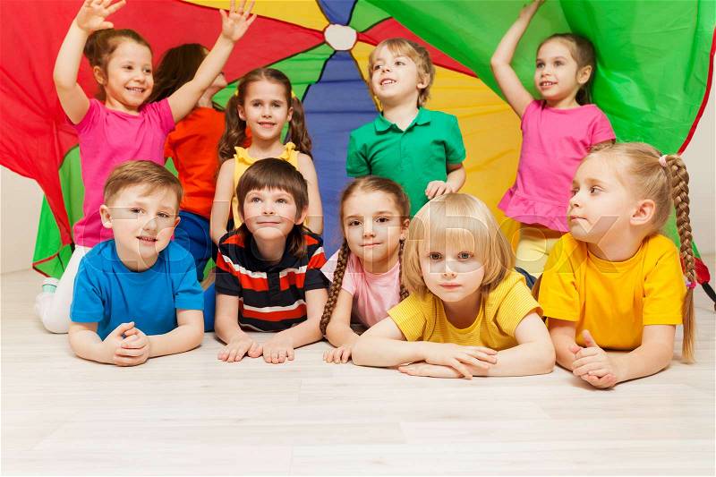 Big group of happy children hiding under canopy made of rainbow parachute during sports games in kindergarten, stock photo