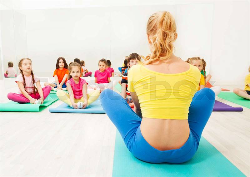 Back view of female gymnastic coach and kid\'s group doing butterfly stretch on yoga mats in exercise room, stock photo