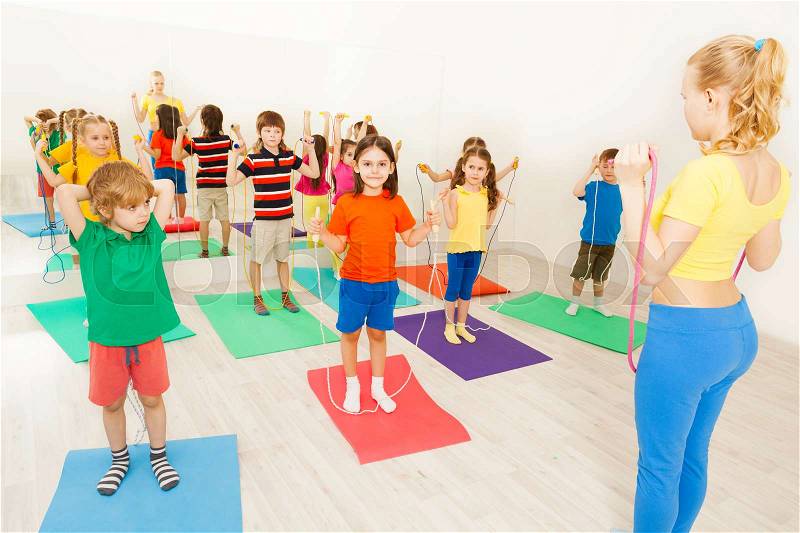 Female gymnastic coach teaching kids doing exercises with jumping rope at sports lesson in gym, stock photo
