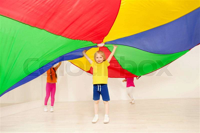 Portrait of blond preschool boy standing under big tent made of colorful parachute in gym, stock photo