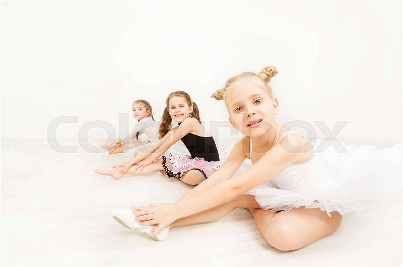 Three little ballerinas, six years old girls in dancewear, sitting on the floor against blanked background of light dance hall, stock photo