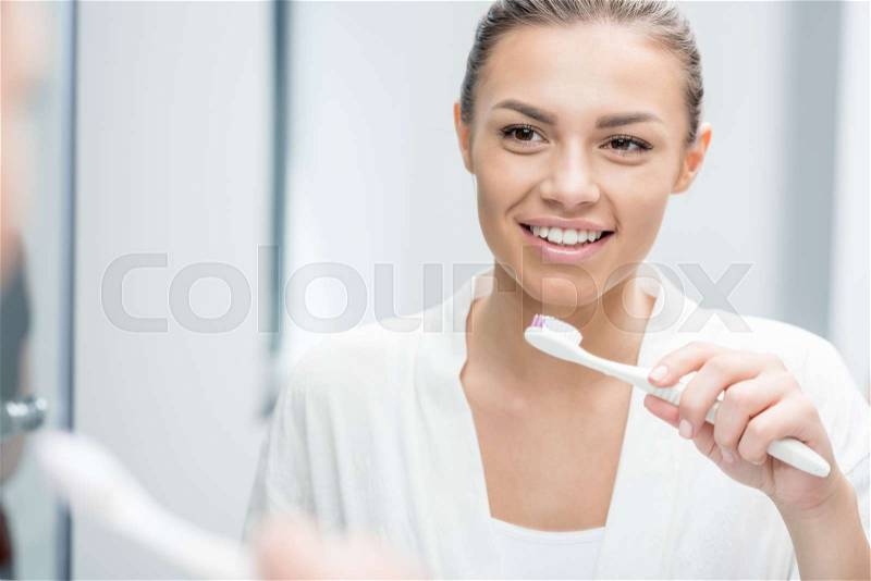 Portrait of happy woman looking in mirror while brushing teeth in morning, stock photo