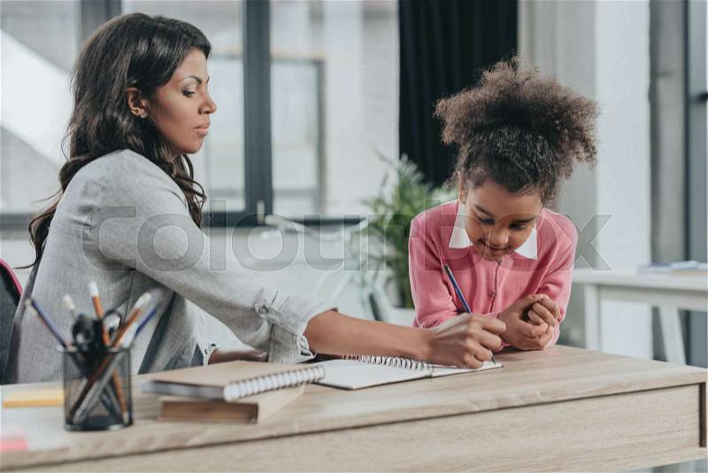 Mother helping little daughter doing homework in business office, stock photo