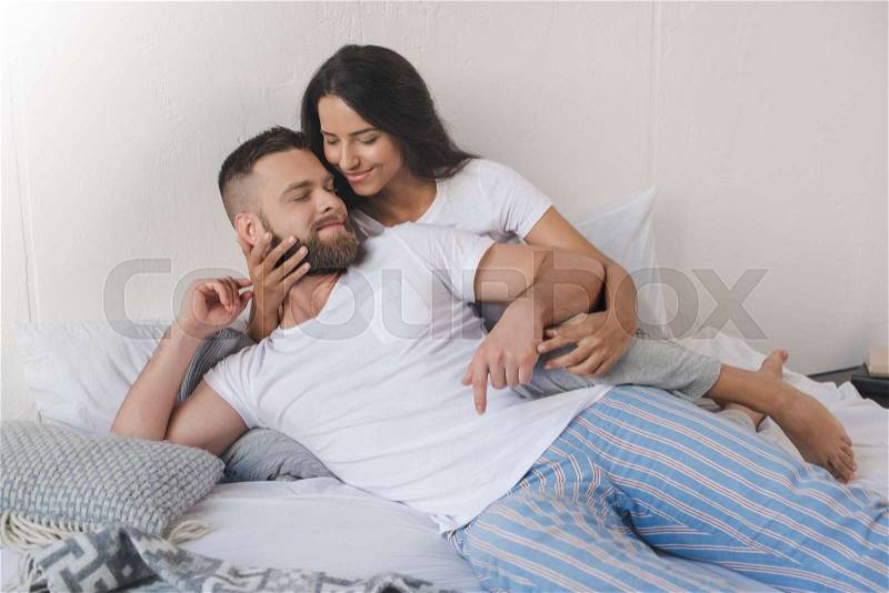 Tender smiling boyfriend and girlfriend hugging and lying on bed at home, stock photo