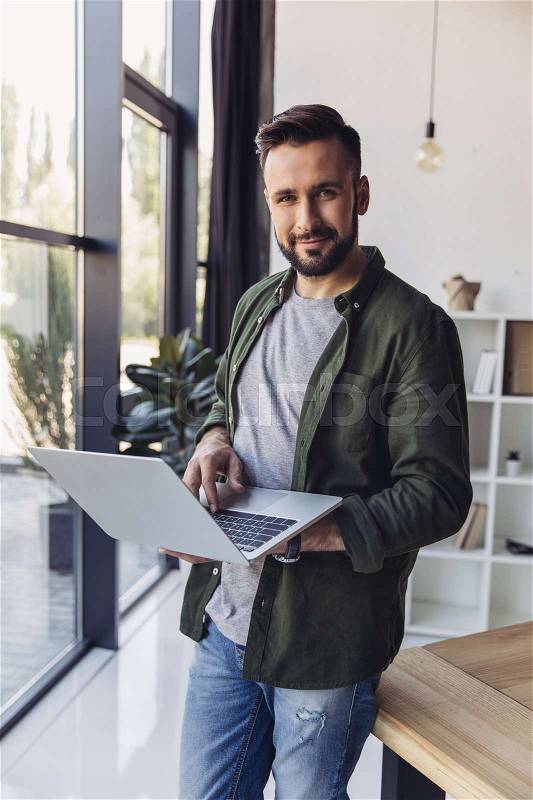 Handsome smiling bearded man using laptop while standing in office, stock photo