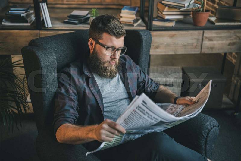 Serious bearded man in eyeglasses reading newspaper while sitting in armchair at home, stock photo