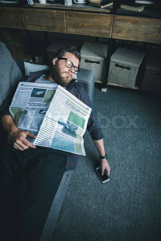 Bearded young man in eyeglasses sleeping with newspaper on sofa at home, stock photo