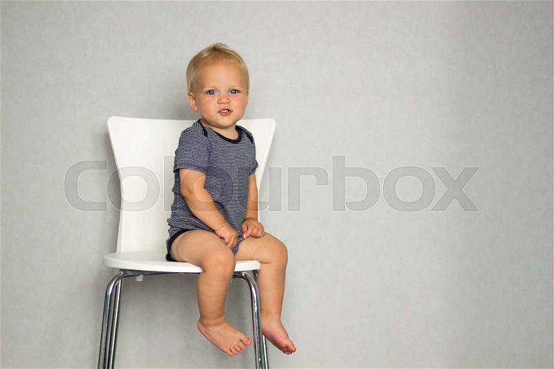 Cute toddler sitting on the chair against gray wall and looking at camera. Copy spase, stock photo