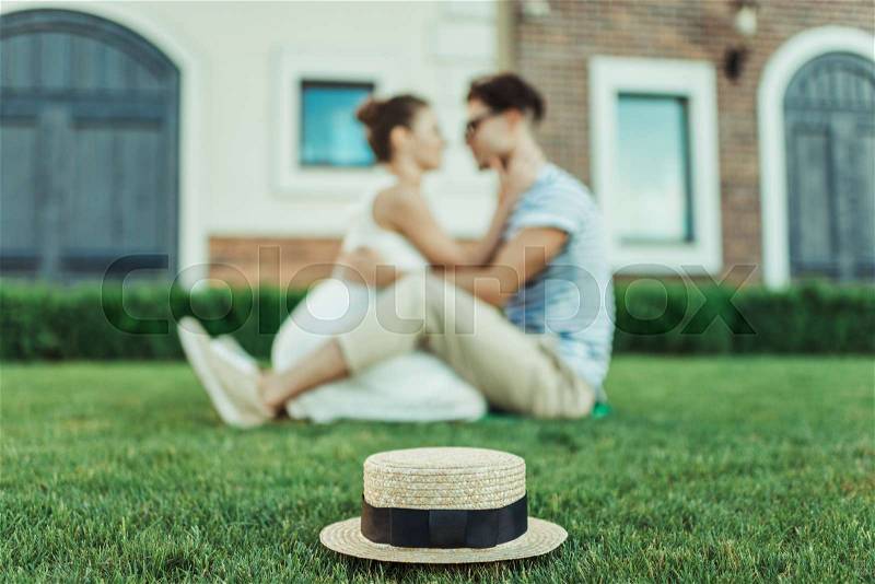 Sweethearts able to kiss while sitting on the grass outdoors, selective focus on straw hat, stock photo