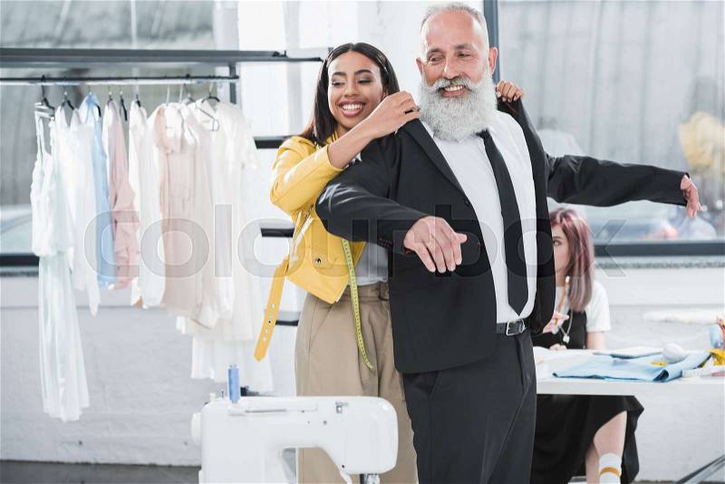 Smiling senior man with young african american stylist fitting suit jacket indoors, stock photo