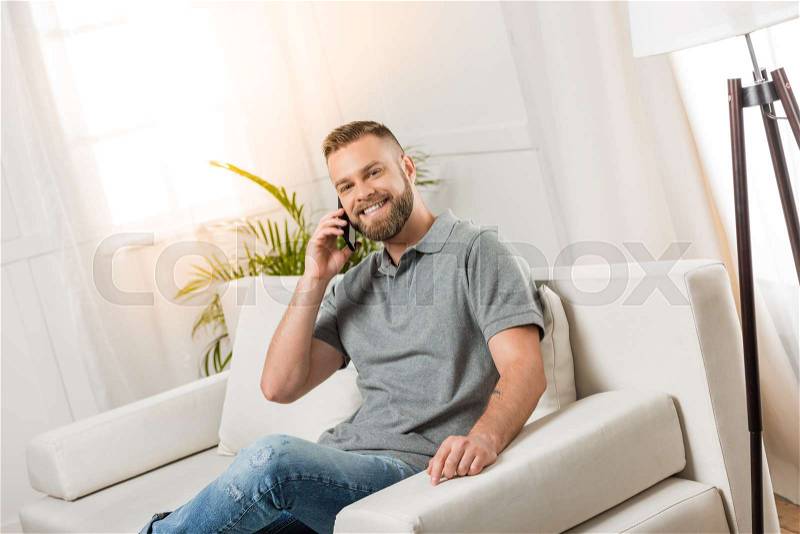 Young smiling man using smartphone and sitting on sofa at home, stock photo