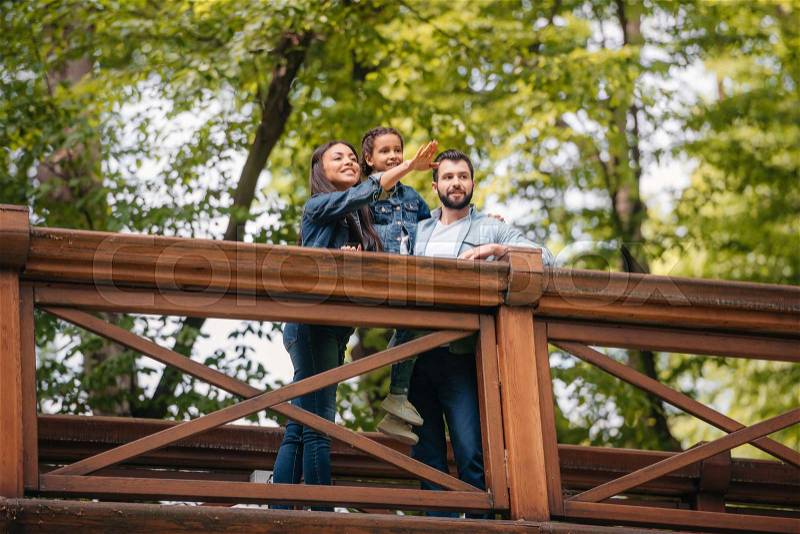 Young happy interracial family standing on wooden bridge, while mother pointing somewhere , stock photo