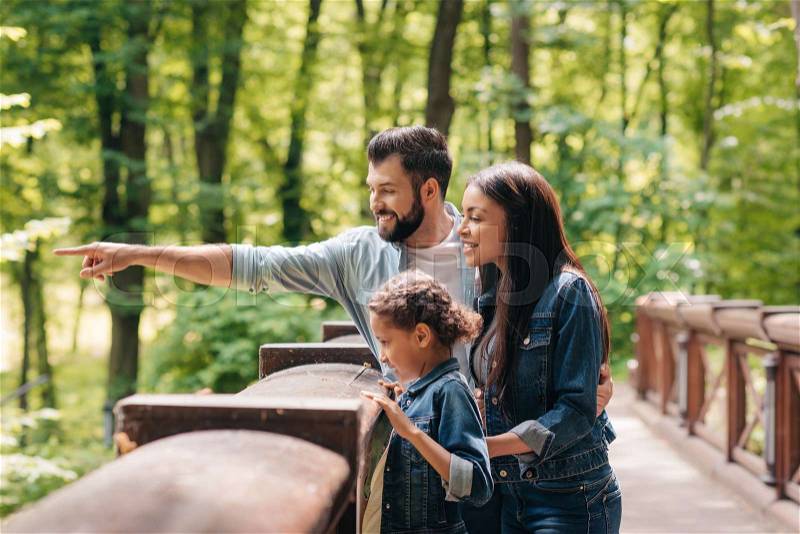 Young smiling interracial family standing on wooden bridge, while father pointing somewhere into the forest, stock photo