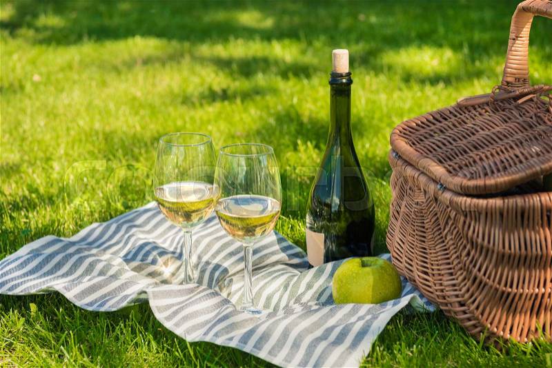 Close-up view of wicker picnic basket, green apple, wine bottle and glasses on napkin in park, stock photo