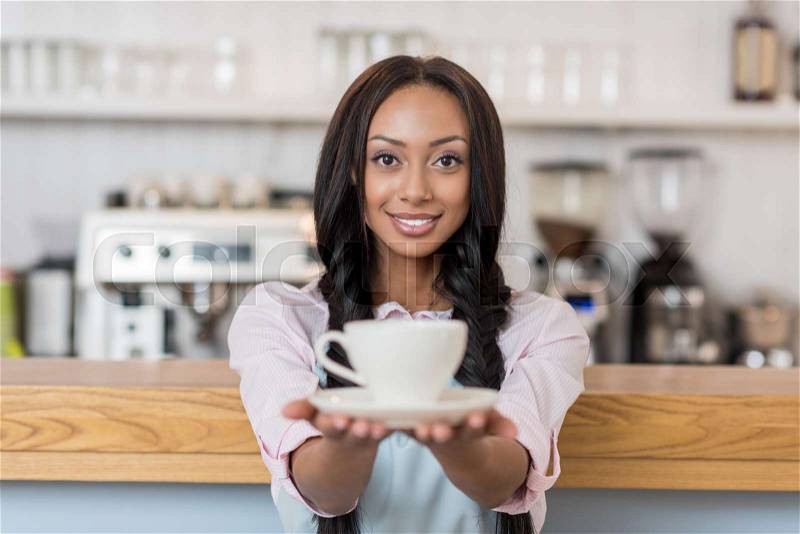 Attractive african american waitress in apron holding coffee cup and looking at camera at cafe, stock photo