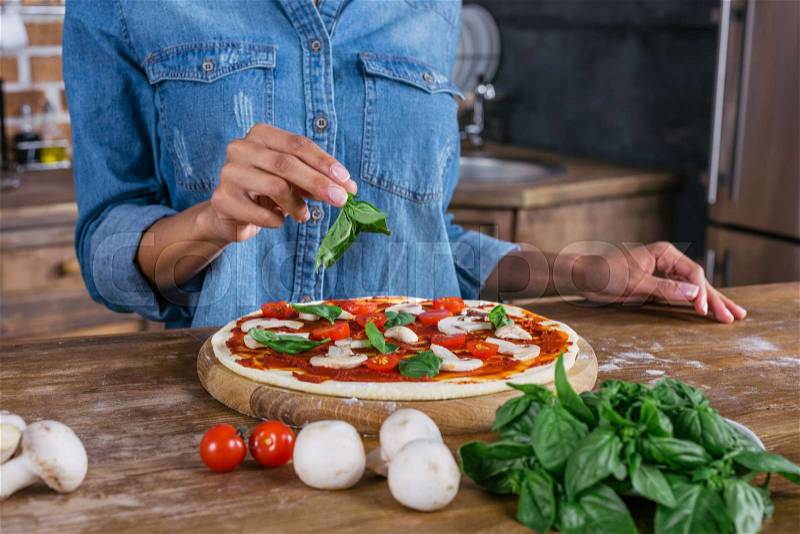 Cropped shot of woman putting basil leaves on raw pizza, stock photo