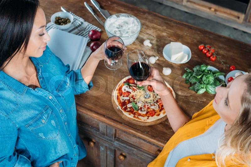 Smiling multiethnic women holding wine glasses while eating pizza at home , stock photo