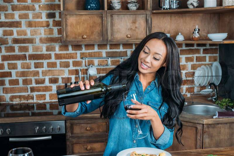 Beautiful smiling african american woman pouring wine in glass at kitchen, stock photo